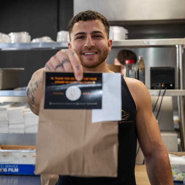 Smiling Holy Bowly employee in a black tank top holding out a brown takeout bag with a sticker.
